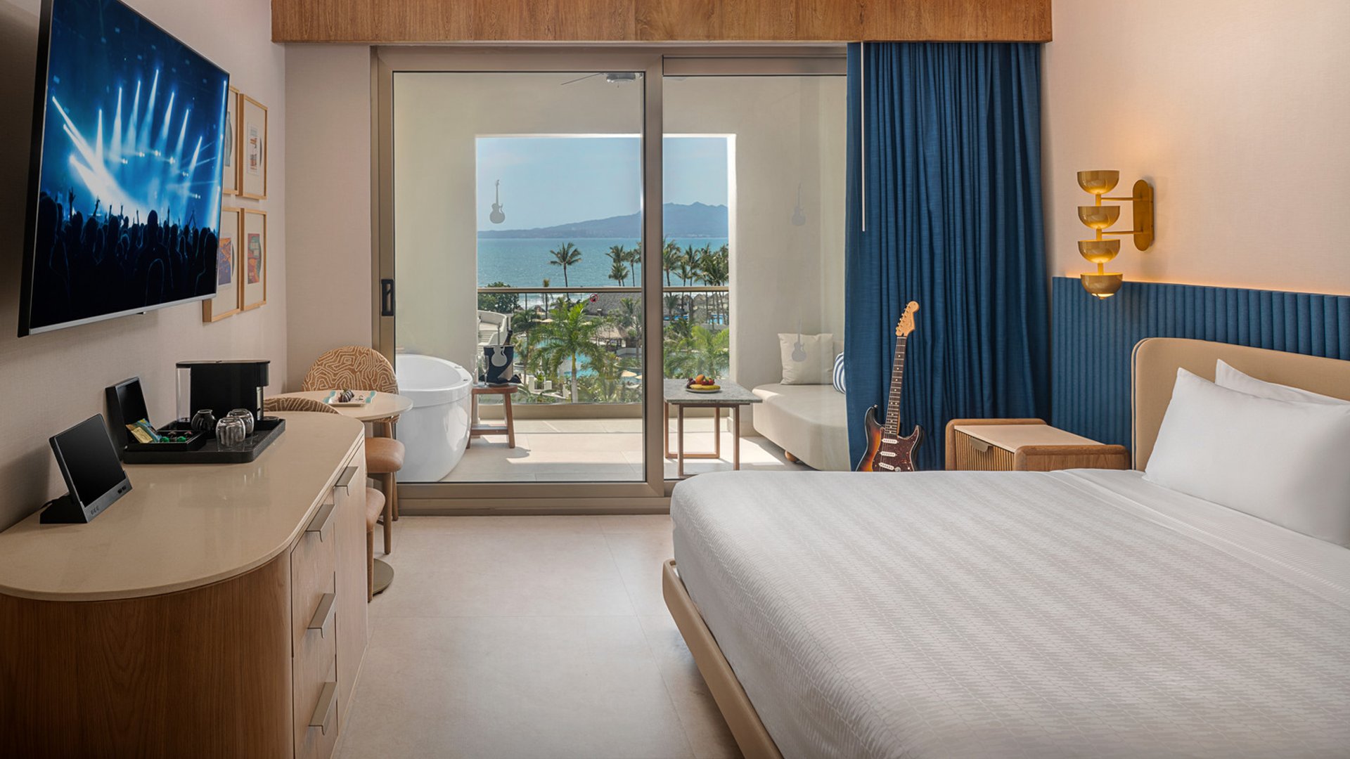 Hotel room with king bed, flatscreen TV, balcony view of ocean and palm trees, electric guitar, and a freestanding bathtub.