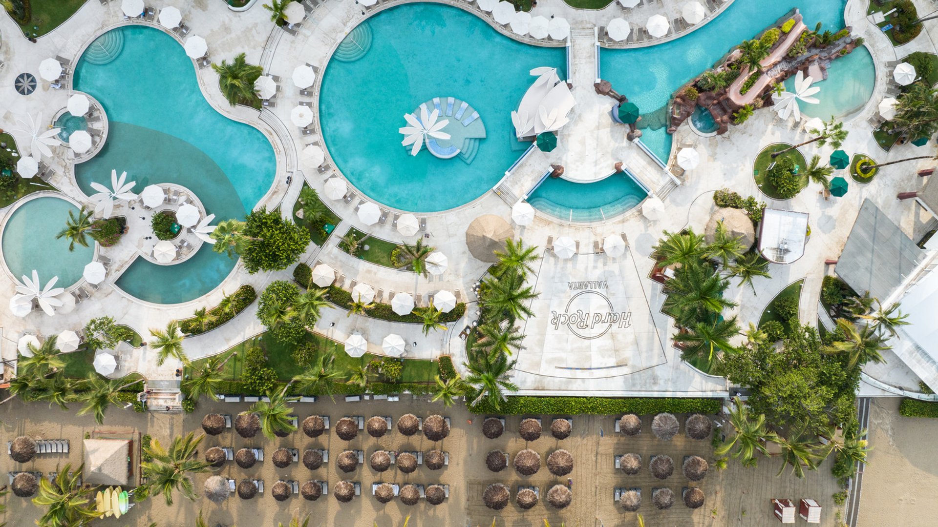 Aerial view of Hard Rock Hotel Vallarta pool area with curvy pools, lounge chairs, and palm trees near the beach.