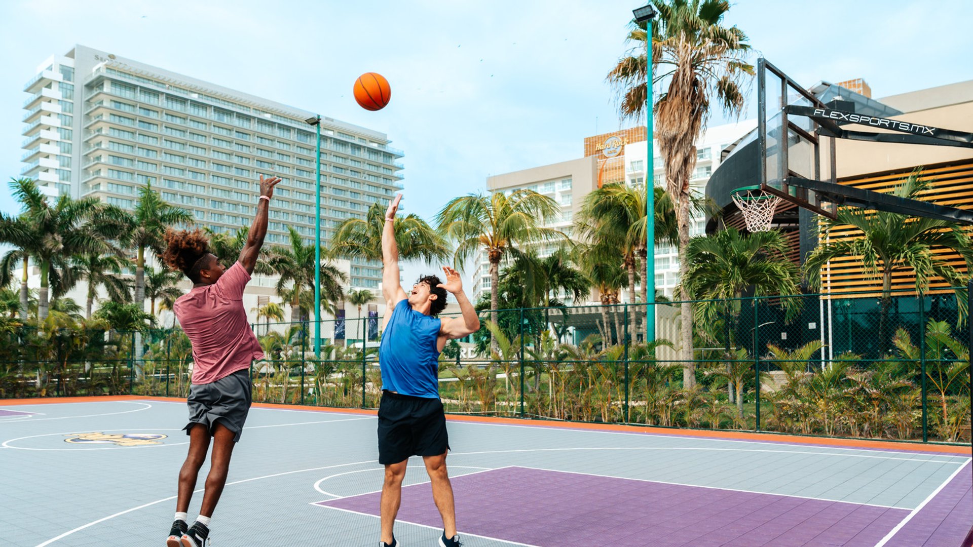Two people playing basketball on an outdoor court at Hard Rock Cancun, surrounded by palm trees and buildings.