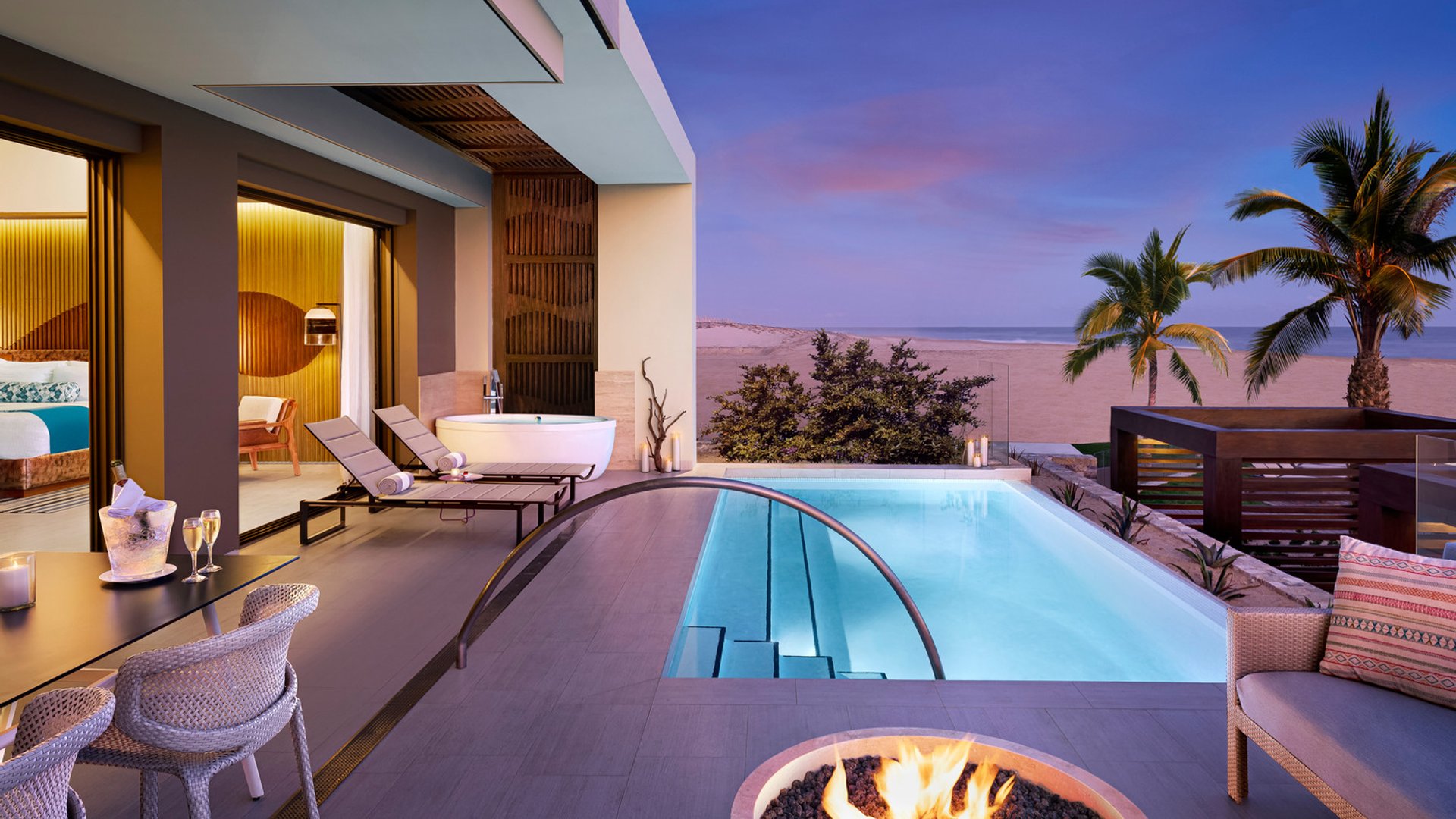 Terrace view with lounge chairs, outdoor bathtub, pool, fire pit, ocean backdrop, and palm trees at dusk.