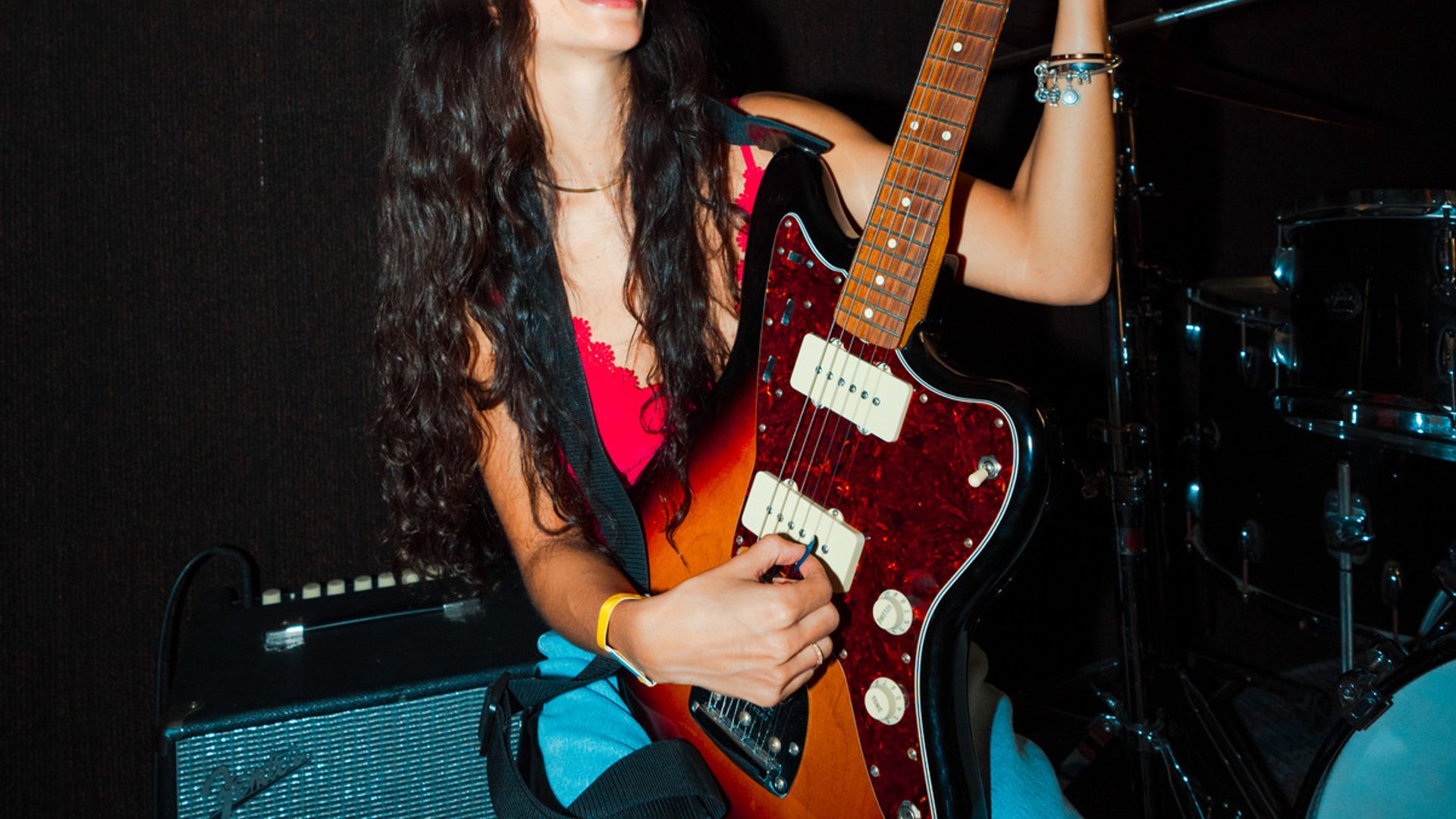 Woman joyfully playing electric guitar in a room with guitars hanging on the wall and a drum set nearby.