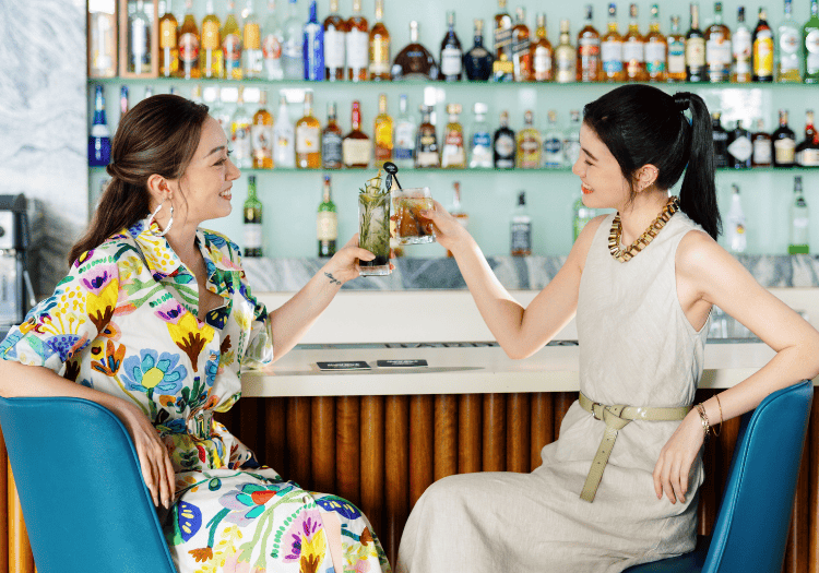 two women sitting at a bar toasting with drinks